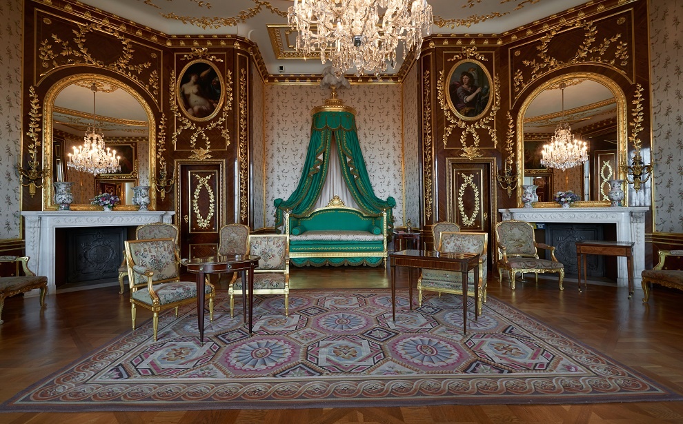 Powiększ obraz: Zdjęcie przedstawia sypialnię króla Stanisława Augusta. Na wprost pod ścianą zielone łoże w kształcie eleganckiej kanapy z rozpiętym u góry baldachimem w tym samym kolorze. Po dwóch stronach łoża marmurowe kominki, nad nimi duże lustra. W nich odbija